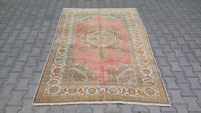 Puede incluir: Una alfombra rectangular vintage con un campo central rosa y dise&ntilde;os de borde intrincados. La alfombra presenta un medall&oacute;n central y detalles en las esquinas en tonos beige, marr&oacute;n y naranja. La alfombra est&aacute; colocada sobre una superficie de ladrillo gris.