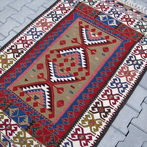 May include: A traditional Turkish kilim rug with a geometric pattern in shades of red, blue, white, and brown. The rug has a fringe border and is woven with wool.