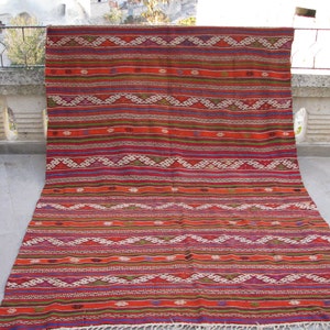 May include: A colorful, handwoven rug with a geometric pattern in shades of red, orange, green, and blue. The rug has a fringe border and is displayed on a stone patio.
