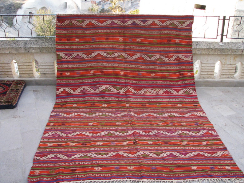 May include: A colorful, handwoven rug with a geometric pattern in shades of red, orange, green, and blue. The rug has a fringe border and is displayed on a stone patio.
