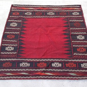 May include: A rectangular, hand-woven rug with a central red field. The rug features a border with geometric patterns in black, white, and red. The rug's dimensions are not visible in the image.