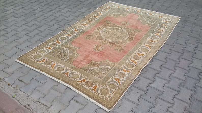 Puede incluir: Alfombra rectangular con un campo central rosa y patrones florales beige y marr&oacute;n ornamentados. El borde presenta un dise&ntilde;o similar con detalles en naranja. La alfombra est&aacute; colocada sobre una superficie de ladrillos grises.