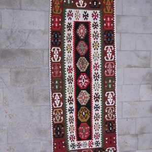 May include: A traditional Turkish rug with a white background and intricate geometric patterns in red, green, brown, and black. The rug has a fringe on the top and bottom.