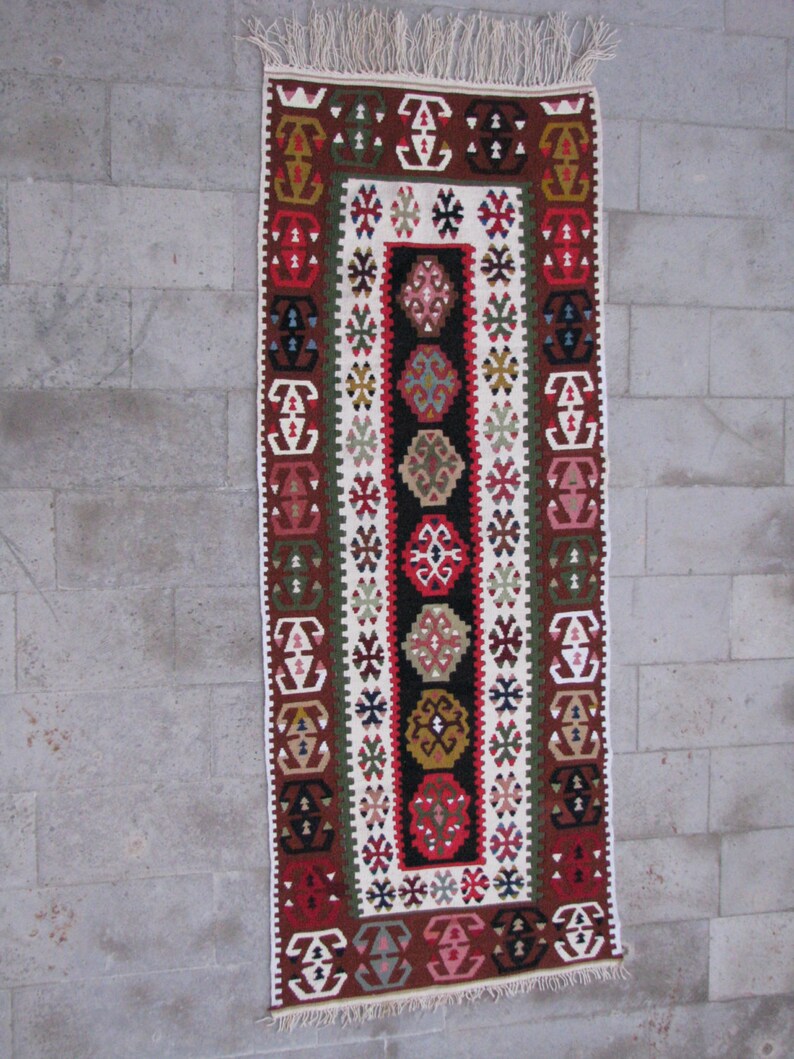 May include: A traditional Turkish rug with a white background and intricate geometric patterns in red, green, brown, and black. The rug has a fringe on the top and bottom.