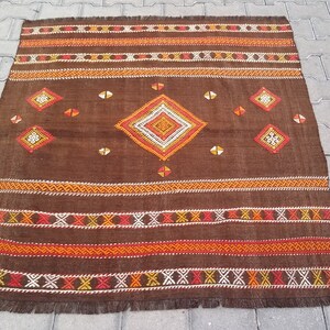May include: A brown wool rug with a geometric pattern in orange, red, and white. The rug has a fringe border.