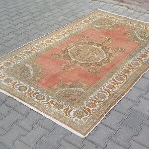 Puede incluir: Alfombra rectangular de estilo vintage con un campo central de color coral y un borde adornado beige y marr&oacute;n. La alfombra presenta un patr&oacute;n sim&eacute;trico detallado en el centro y a lo largo de los bordes, creando un aspecto cl&aacute;sico y elegante. La alfombra est&aacute; colocada sobre una superficie de ladrillos grises.