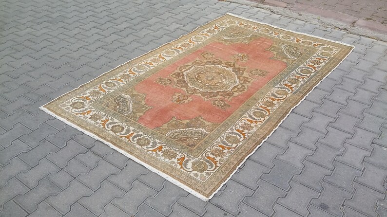 Puede incluir: Alfombra rectangular de estilo vintage con un campo central de color coral y un borde adornado beige y marr&oacute;n. La alfombra presenta un patr&oacute;n sim&eacute;trico detallado en el centro y a lo largo de los bordes, creando un aspecto cl&aacute;sico y elegante. La alfombra est&aacute; colocada sobre una superficie de ladrillos grises.