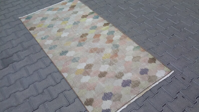 May include: A rectangular rug with a repeating pattern of colorful, cloud-like shapes in pastel shades of pink, yellow, blue, and brown. The rug has a beige background and a fringed edge. The rug is placed on a gray brick surface.