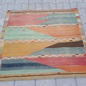 May include: A vintage rug with a geometric pattern in shades of orange, red, blue, and green. The rug has a fringe border and is made of wool.