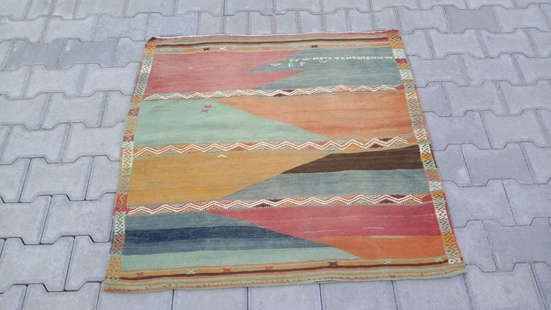 May include: A vintage rug with a geometric pattern in shades of orange, red, blue, and green. The rug has a fringe border and is made of wool.