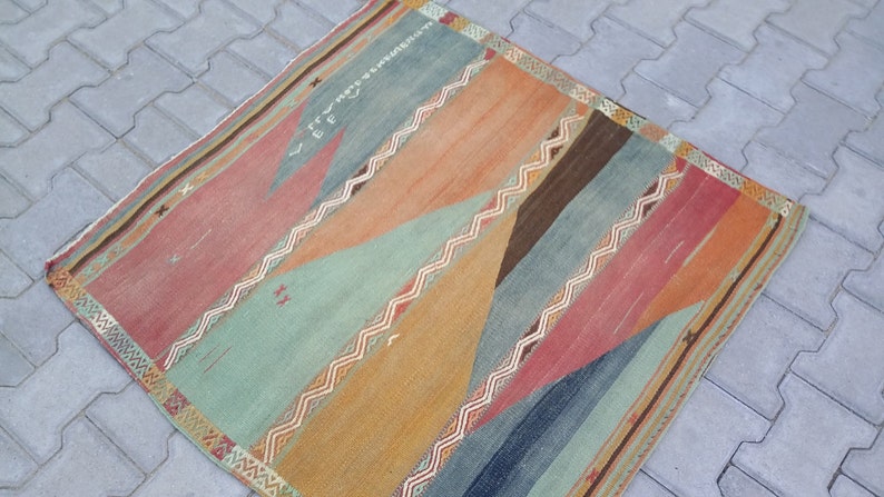May include: A colorful, geometric patterned rug with a mix of red, orange, blue, green, and brown tones. The rug has a fringed border.
