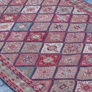 May include: A vintage rug with a diamond pattern in shades of red, blue, and beige. The rug has a detailed floral design and a border with a repeating pattern.