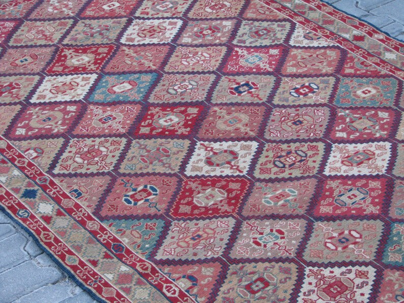 May include: A vintage rug with a diamond pattern in shades of red, blue, and beige. The rug has a detailed floral design and a border with a repeating pattern.