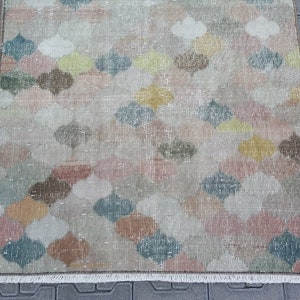 May include: A rectangular rug with a repeating pattern of colorful, stylized floral shapes in shades of pink, yellow, green, and brown. The rug has a distressed, vintage look with a fringed edge. The rug is placed on a gray surface.