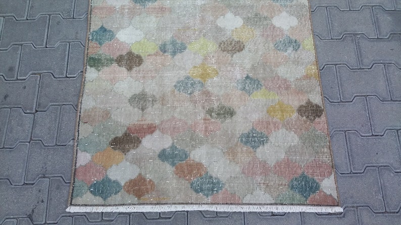 May include: A rectangular rug with a repeating pattern of colorful, stylized floral shapes in shades of pink, yellow, green, and brown. The rug has a distressed, vintage look with a fringed edge. The rug is placed on a gray surface.