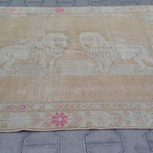 以下が含まれることがあります： ベージュの背景に、向かい合った2匹のライオンの薄れたデザインが施されたヴィンテージラグ。ラグはピンクと白のボーダーで、幾何学模様が描かれている。