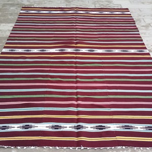 May include: A striped woven rug with a burgundy background and colorful stripes in shades of green, blue, pink, and yellow. The rug has a simple geometric pattern in the center.