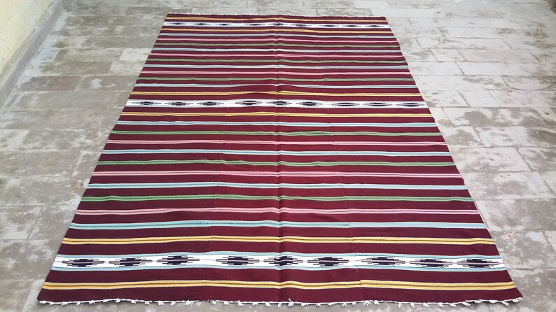 May include: A striped woven rug with a burgundy background and colorful stripes in shades of green, blue, pink, and yellow. The rug has a simple geometric pattern in the center.
