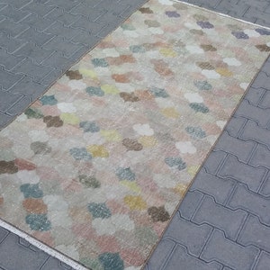 May include: A rectangular rug with a repeating pattern of cloud-like shapes in pastel colors including pink, yellow, blue, and brown. The rug has a cream-colored background and a fringed edge. The rug is placed on a gray brick surface.
