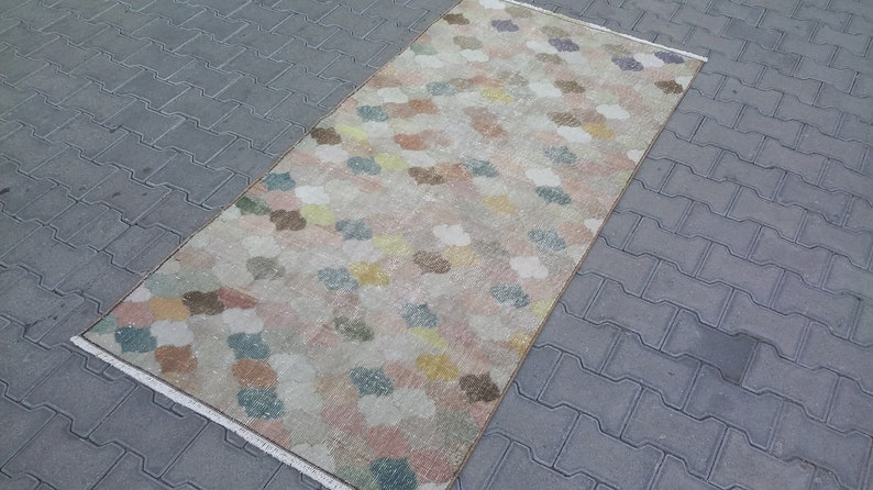 May include: A rectangular rug with a repeating pattern of cloud-like shapes in pastel colors including pink, yellow, blue, and brown. The rug has a cream-colored background and a fringed edge. The rug is placed on a gray brick surface.