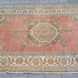 Puede incluir: Una alfombra rectangular de estilo vintage con un dise&ntilde;o de medall&oacute;n central. La alfombra presenta un campo rosa descolorido, bordeado por un patr&oacute;n crema y verde oliva con motivos florales y geom&eacute;tricos. Los bordes de la alfombra est&aacute;n deshilachados.