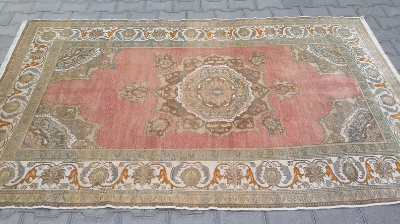 Puede incluir: Una alfombra rectangular de estilo vintage con un dise&ntilde;o de medall&oacute;n central. La alfombra presenta un campo rosa descolorido, bordeado por un patr&oacute;n crema y verde oliva con motivos florales y geom&eacute;tricos. Los bordes de la alfombra est&aacute;n deshilachados.
