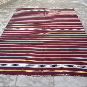 May include: A striped woven rug with a burgundy background and colorful stripes in shades of pink, green, yellow, and white. The rug has a fringe border.