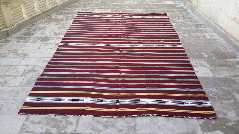 May include: A striped woven rug with a burgundy background and colorful stripes in shades of pink, green, yellow, and white. The rug has a fringe border.