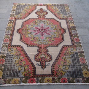 Puede incluir: Una alfombra turca vintage con un diseño central de medallón en tonos de rosa, rojo, amarillo y gris. La alfombra tiene un fondo crema y un borde con flecos.