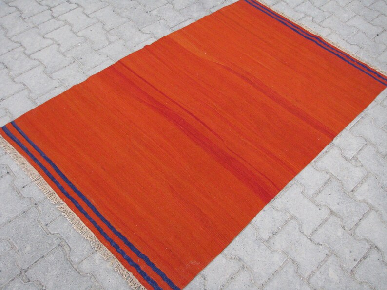 K&ouml;nnte beinhalten: Ein leuchtend orangefarbener Teppich mit zwei dunkelblauen Streifen an den R&auml;ndern. Der Teppich hat einen Fransenrand.