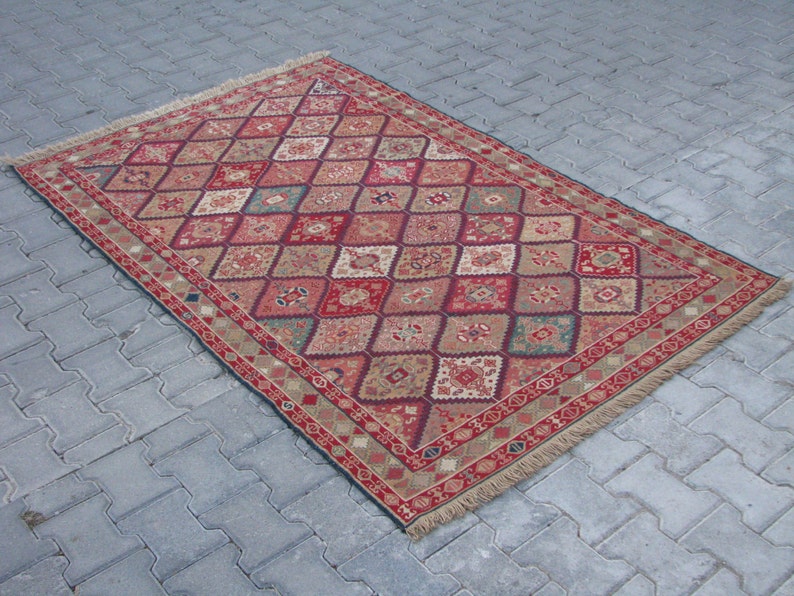 May include: A red, beige, and green patterned rug with a diamond design. The rug has a fringe border.