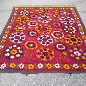 Puede incluir: Un gran tapiz de pared rectangular de tela roja con un dise&ntilde;o bordado decorativo. El dise&ntilde;o presenta una variedad de flores coloridas y c&iacute;rculos en tonos de blanco, amarillo, rosa, morado y verde.
