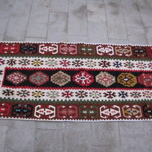May include: A traditional Turkish rug with a white background and intricate geometric patterns in red, blue, green, and brown. The rug has a central band with a repeating pattern of stylized flowers and leaves.
