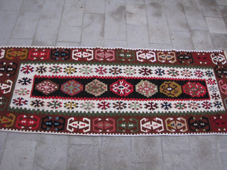 May include: A traditional Turkish rug with a white background and intricate geometric patterns in red, blue, green, and brown. The rug has a central band with a repeating pattern of stylized flowers and leaves.