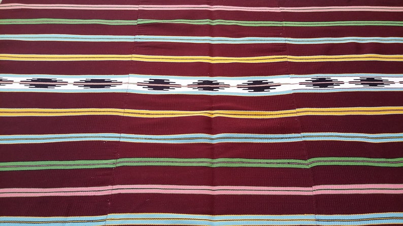 May include: A woven fabric with a striped pattern in shades of red, yellow, green, blue, and white. The fabric has a geometric design in the center.
