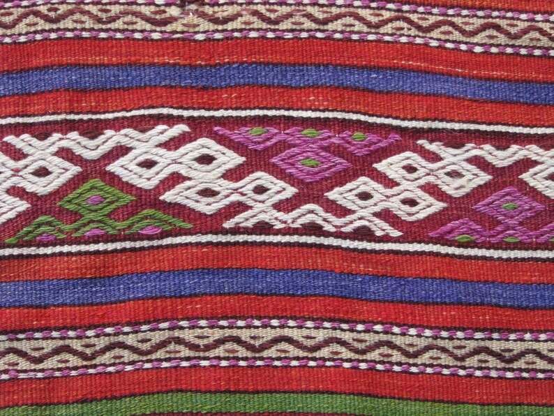 May include: A close-up of a woven fabric with a repeating pattern of red, blue, white, green, and purple. The fabric has a textured surface and a traditional design.