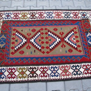 May include: A traditional Turkish rug with a red, blue, and white geometric pattern. The rug has a fringe on the ends and is made of wool.