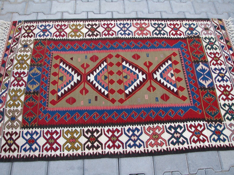 May include: A traditional Turkish rug with a red, blue, and white geometric pattern. The rug has a fringe on the ends and is made of wool.