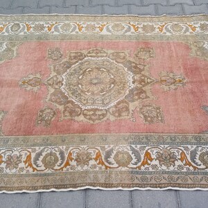 Puede incluir: Una alfombra rectangular de estilo vintage con un campo central rosa y un borde floral ornamentado. La alfombra presenta un dise&ntilde;o detallado y sim&eacute;trico en tonos beige, marr&oacute;n y naranja. Los bordes de la alfombra est&aacute;n deshilachados.