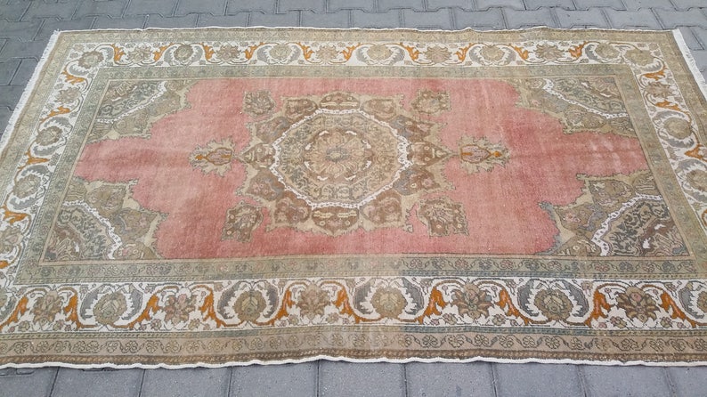 Puede incluir: Una alfombra rectangular de estilo vintage con un campo central rosa y un borde floral ornamentado. La alfombra presenta un dise&ntilde;o detallado y sim&eacute;trico en tonos beige, marr&oacute;n y naranja. Los bordes de la alfombra est&aacute;n deshilachados.