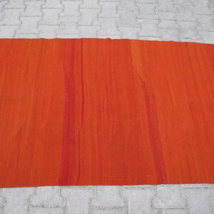 K&ouml;nnte beinhalten: Ein gro&szlig;er, rechteckiger, orangefarbener Teppich mit einem blauen und wei&szlig;en Rand. Der Teppich hat eine gewebte Textur und ein subtiles Streifenmuster.