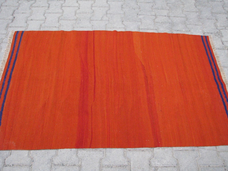 K&ouml;nnte beinhalten: Ein gro&szlig;er, rechteckiger, orangefarbener Teppich mit einem blauen und wei&szlig;en Rand. Der Teppich hat eine gewebte Textur und ein subtiles Streifenmuster.