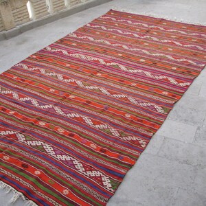May include: A colorful, handwoven rug with a geometric pattern in shades of red, orange, green, blue, and white. The rug has a fringe border and is made of wool.