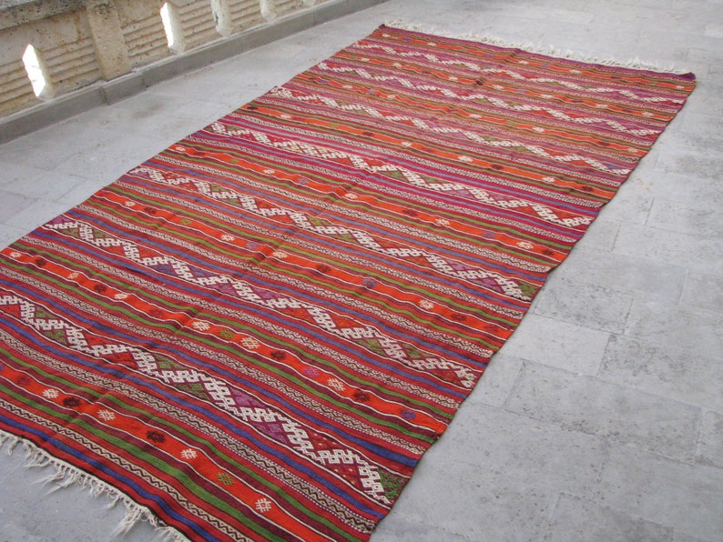May include: A colorful, handwoven rug with a geometric pattern in shades of red, orange, green, blue, and white. The rug has a fringe border and is made of wool.