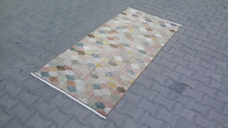 May include: Rectangular rug with a repeating pattern of colorful, cloud-like shapes in pastel shades of pink, yellow, blue, and brown. The rug has a cream-colored border and is placed on a gray, textured surface.