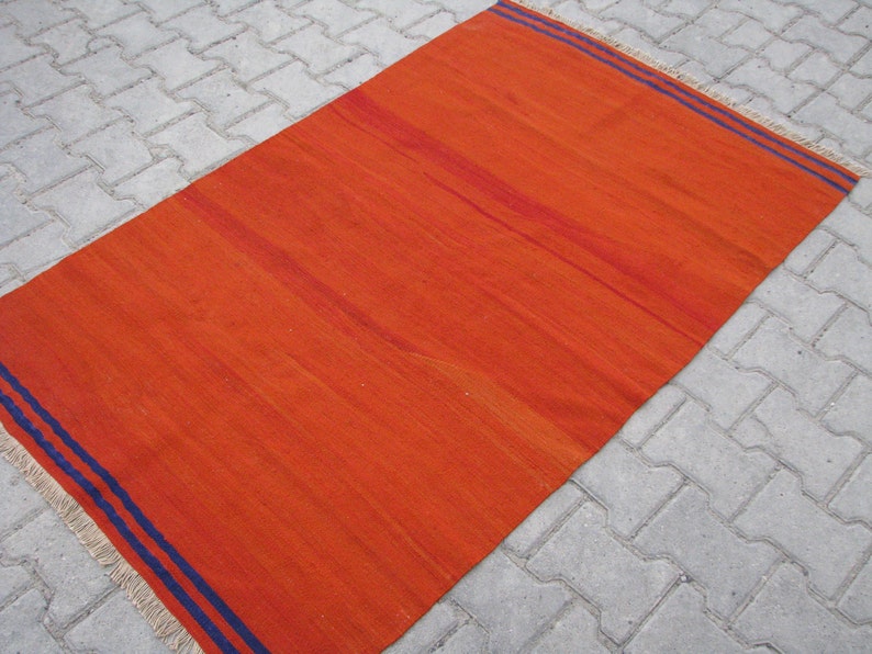 K&ouml;nnte beinhalten: Ein gro&szlig;er, rechteckiger, orangefarbener Teppich mit zwei blauen Streifen an den R&auml;ndern. Der Teppich hat einen Fransenrand.