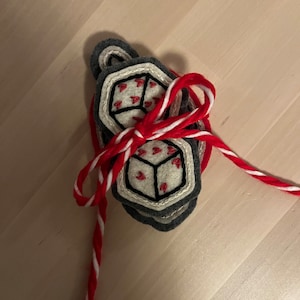 May include: A set of three felt dice ornaments with red hearts embroidered on each face. The dice are tied together with a red and white striped twine.