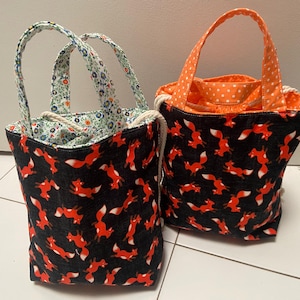 May include: Two tote bags. One bag has a black background with orange foxes, floral lining, and floral handles. The other bag has an orange polka dot lining and handles, with a black background and orange foxes.