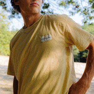 May include: A young person wearing a yellow t-shirt with a small patch that says "michigan" on the left side of the chest. The person is standing in a forest with trees in the background.