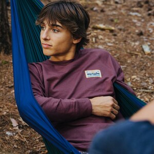 May include: A young person wearing a maroon long-sleeved shirt with a patch that says "michigan" in white lettering on a blue background. The person is sitting in a blue and green hammock.